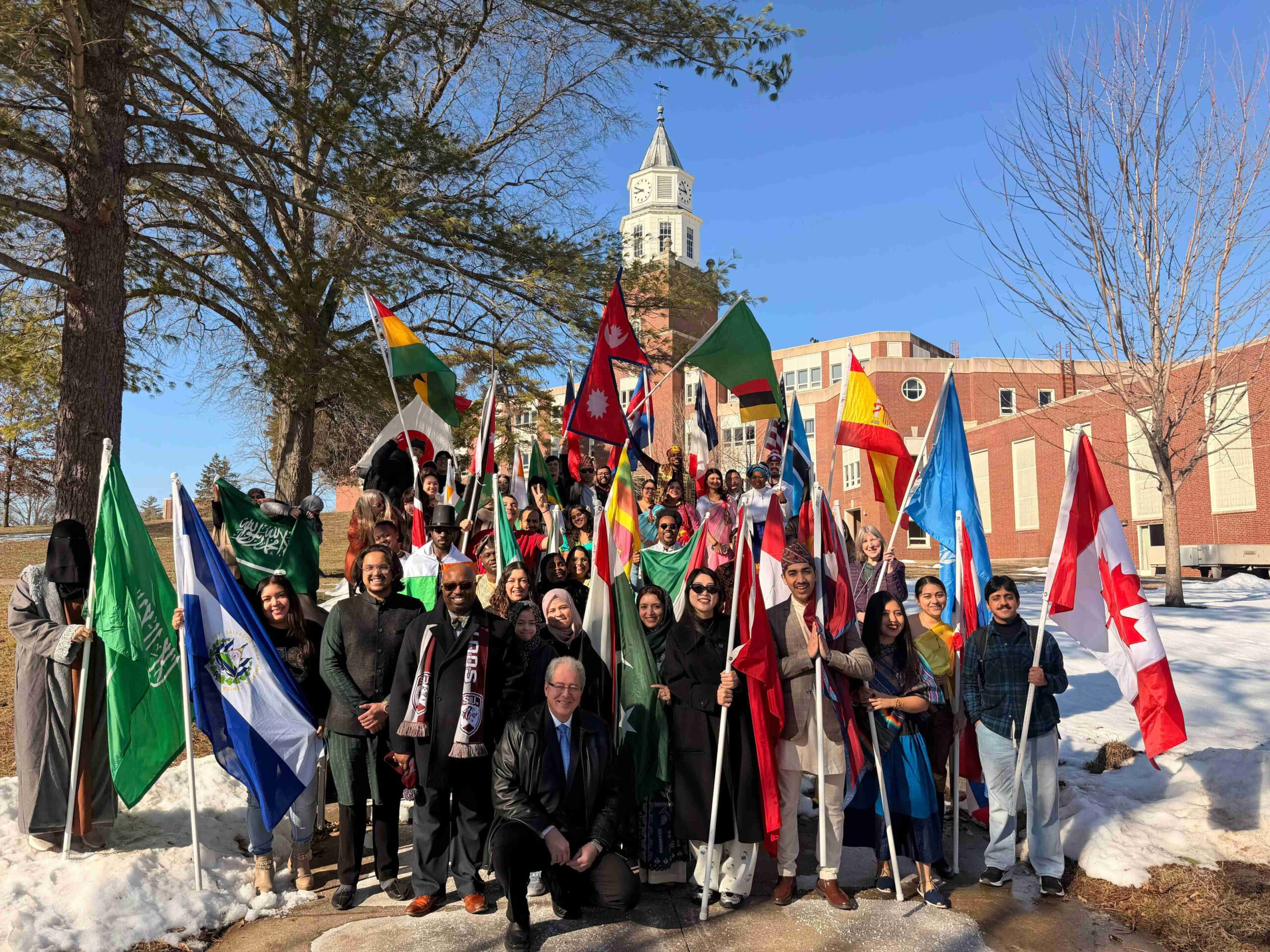 International student and faculty members stand in front of the SIU Carbondale clocktower holding national flags from multiple countries during the International Flag Parade 2026. Snow is visible on the ground, and campus buildings and trees appear in the background.
