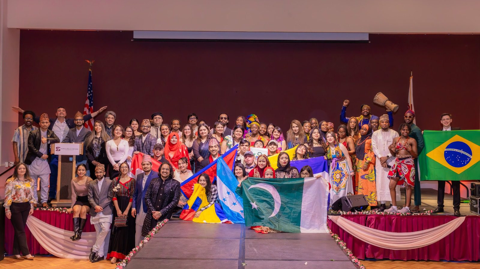 International students gather on stage during the International Cultural Night 2026, wearing traditional clothing and holding national flags from different countries for a group photo.